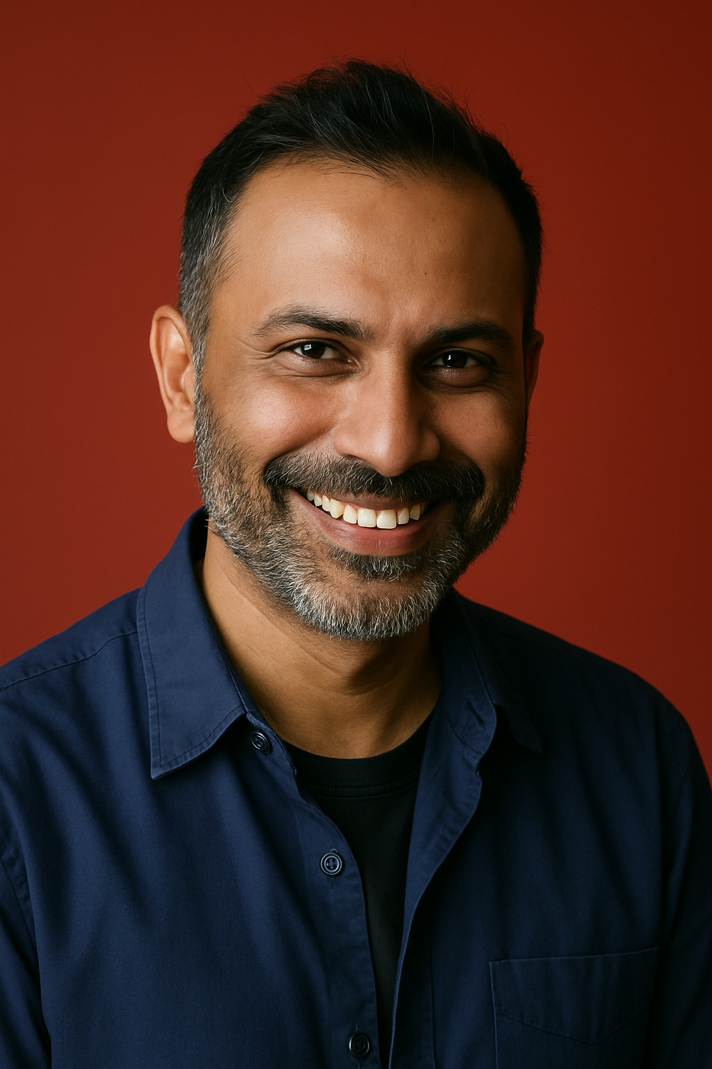A close-up portrait of a man with short hair and a salt-and-pepper beard, smiling warmly while wearing a navy blue shirt in front of a red background.