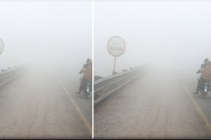 Motorcyclists riding slowly on a fog-covered North India road with very low visibility and a ‘SLOW’ sign barely visible.